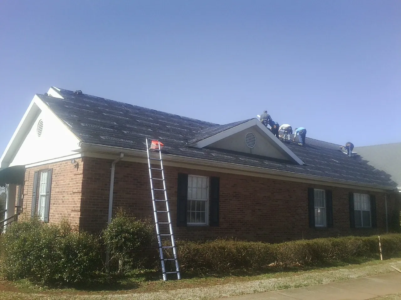 Crew tearing shingles off a brick ranch roof with a ladder set — Emergency Roof Repair work by our crew in Bells