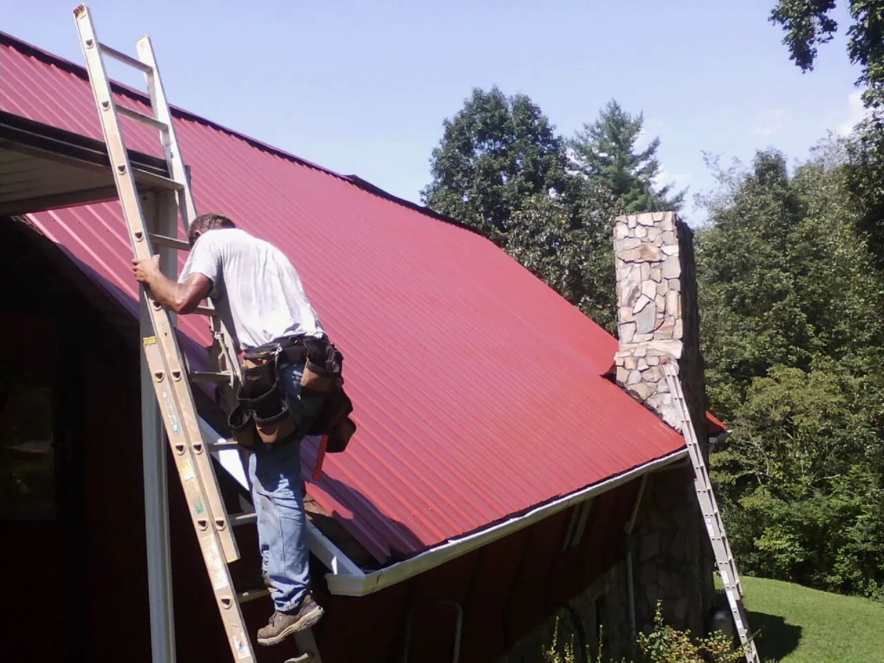 Roofer on a ladder beside a red metal roof and stone chimney — Soffit & Fascia Repair by Roofing Brothers in Bells